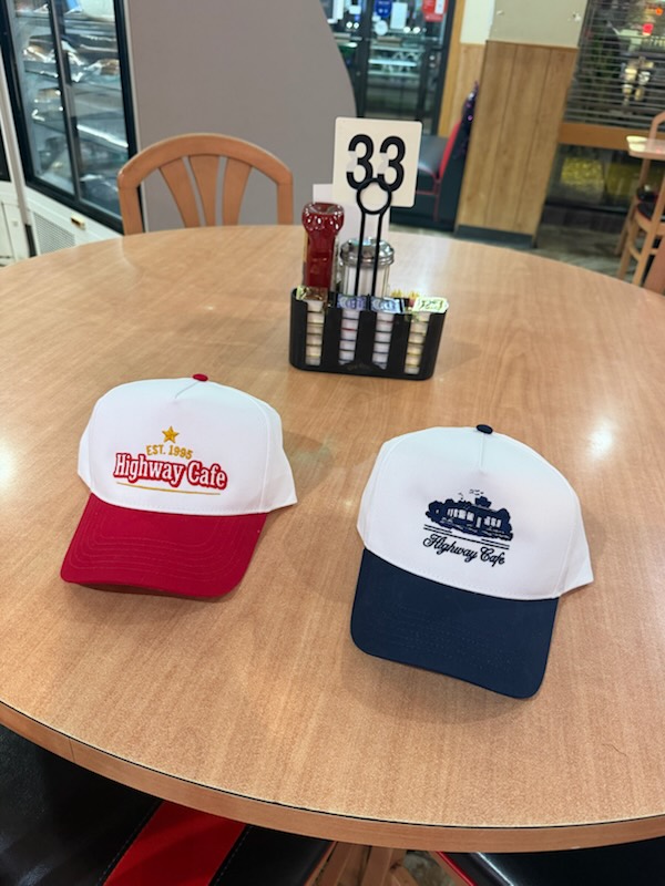 Two Highway Cafe snapback hats on a diner table: one red and white, one navy and white